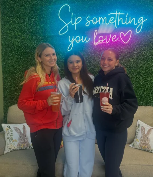 Three customers with drinks in front of the Sip Something You Love neon sign at Restore Nutrition