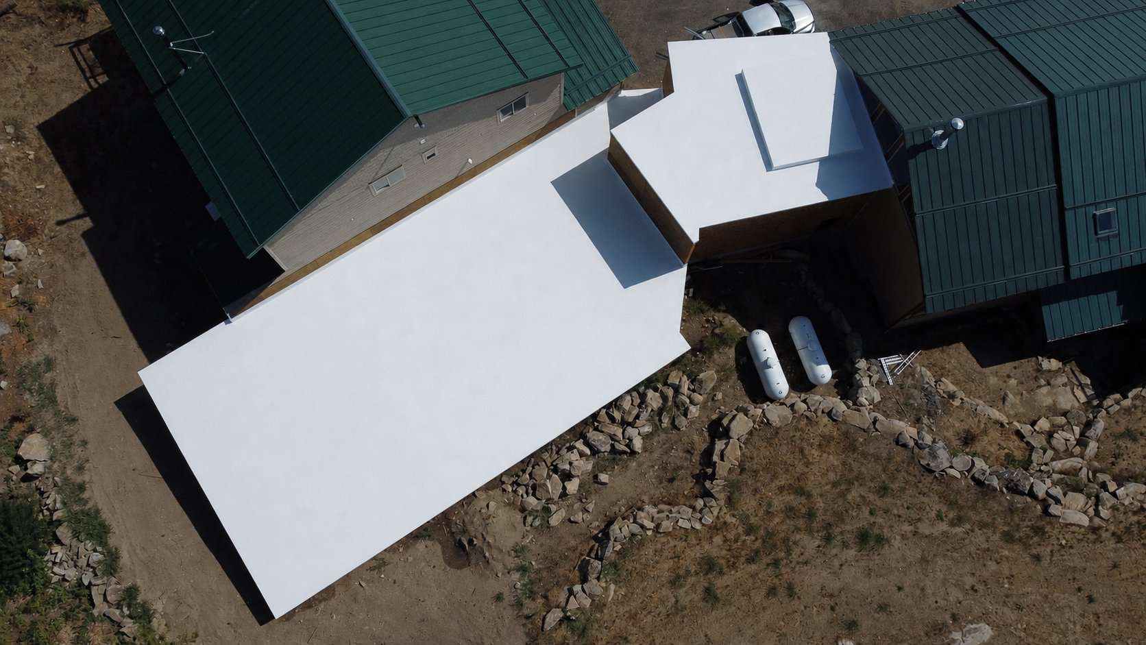 Aerial drone view of a completed white flat roof membrane installation beside metal-roofed outbuildings.