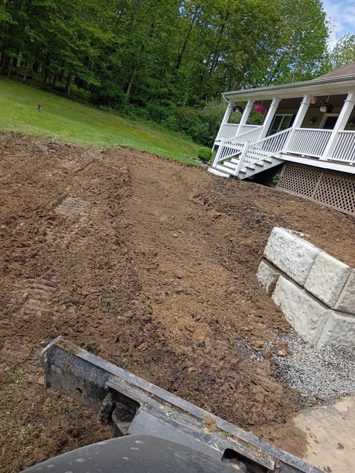 Grading and soil preparation work in progress on a residential property sloped landscape with a white cottage-style house in the background.
