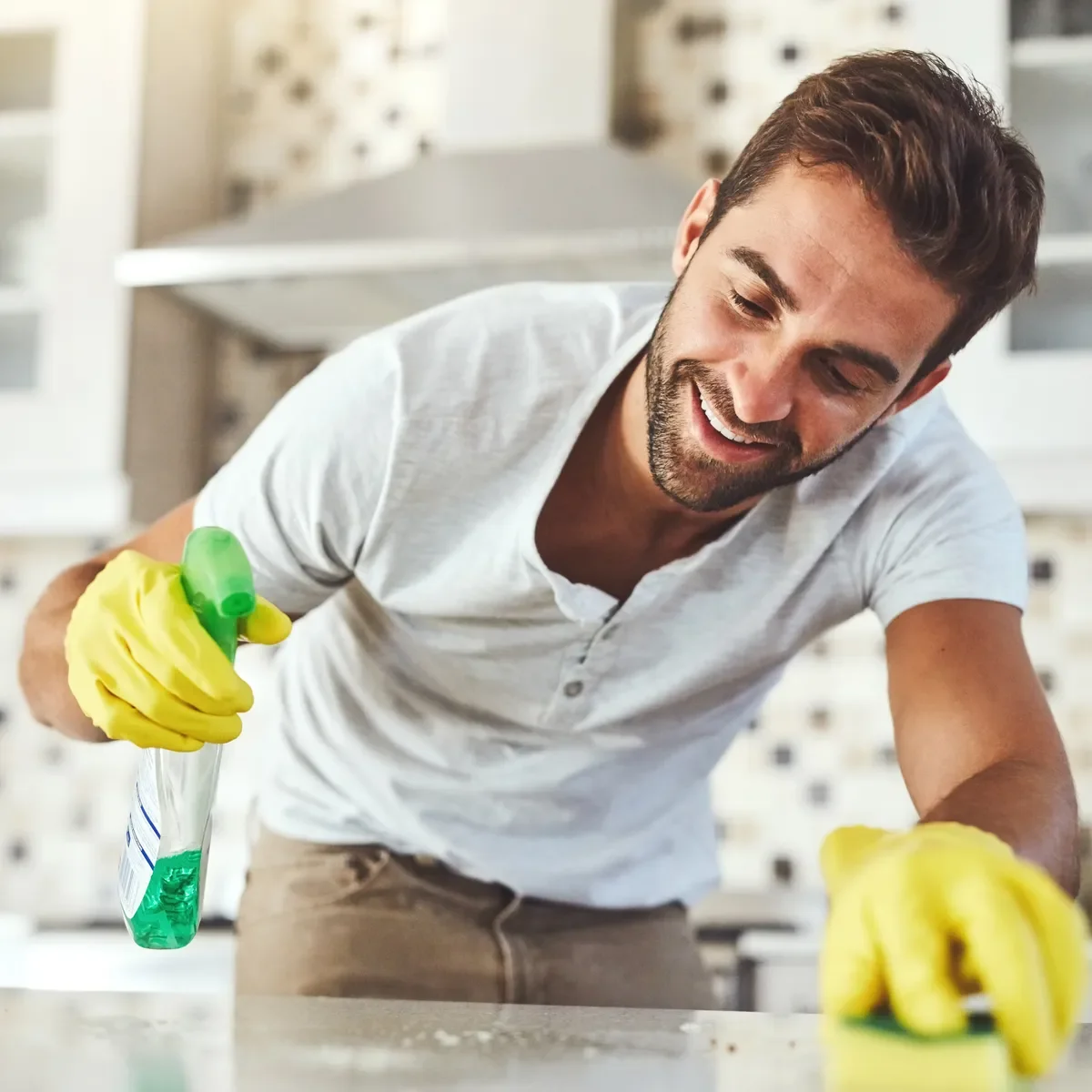 A man in yellow gloves spraying cleaning solution on a kitchen countertop, demonstrating surface cleaning work in a modern kitchen interior.