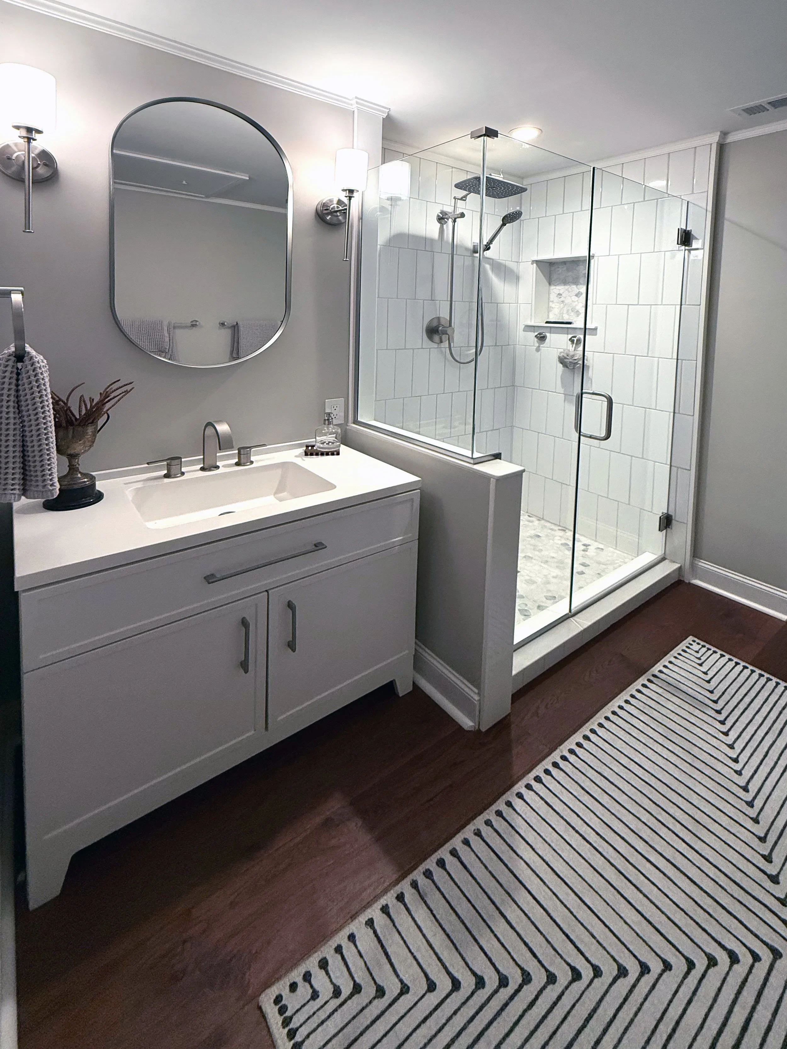 A complete bathroom remodel featuring a white vanity with sink, framed mirror, and a large walk-in shower with white subway tile and glass enclosure in a modern residential bathroom.