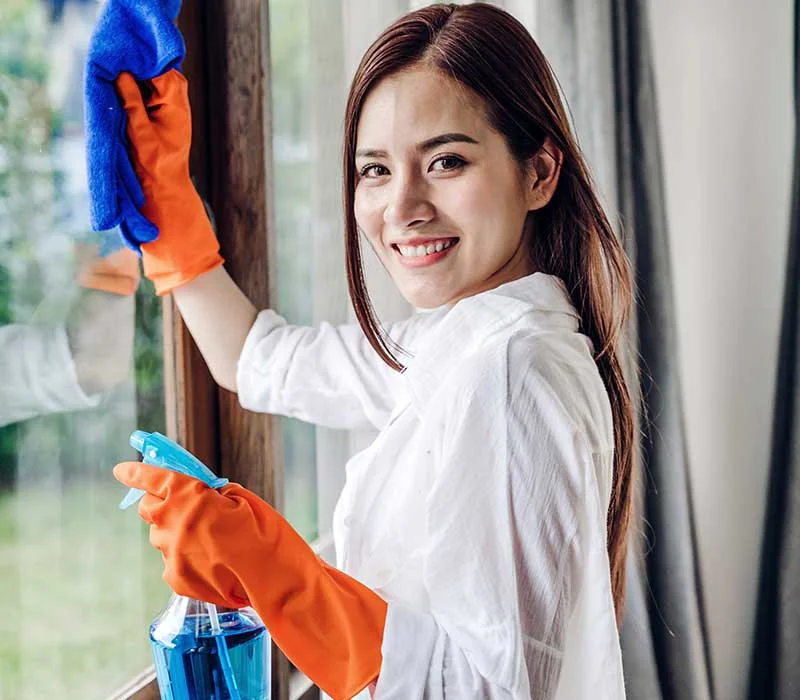 A cleaning service worker wearing protective gloves and using cleaning supplies to wash a window in an interior setting.