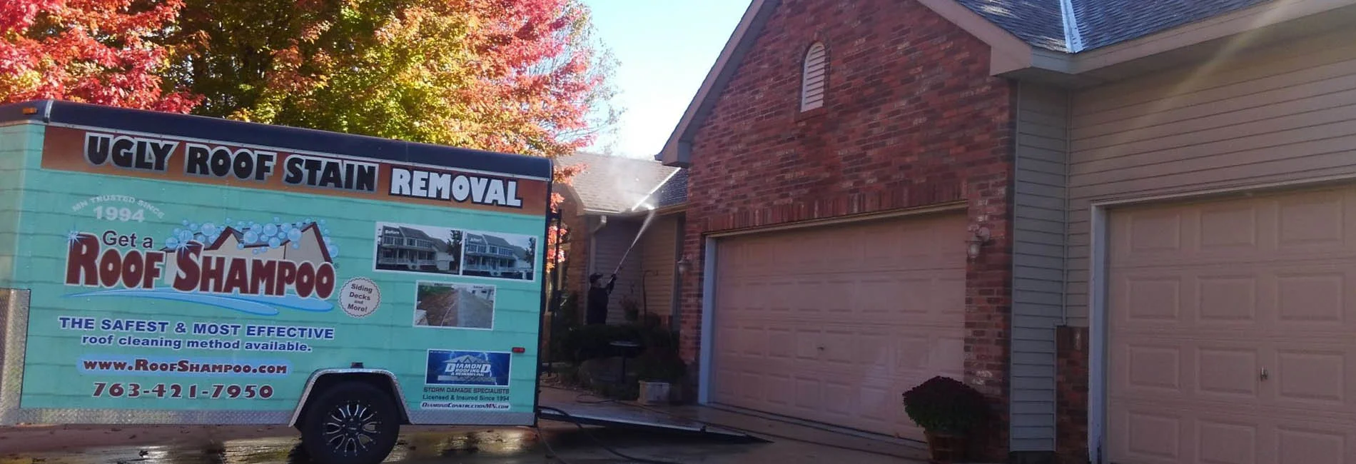 A roof cleaning service trailer advertising 'Roof Shampoo' stain removal parked in a residential driveway next to a brick house.