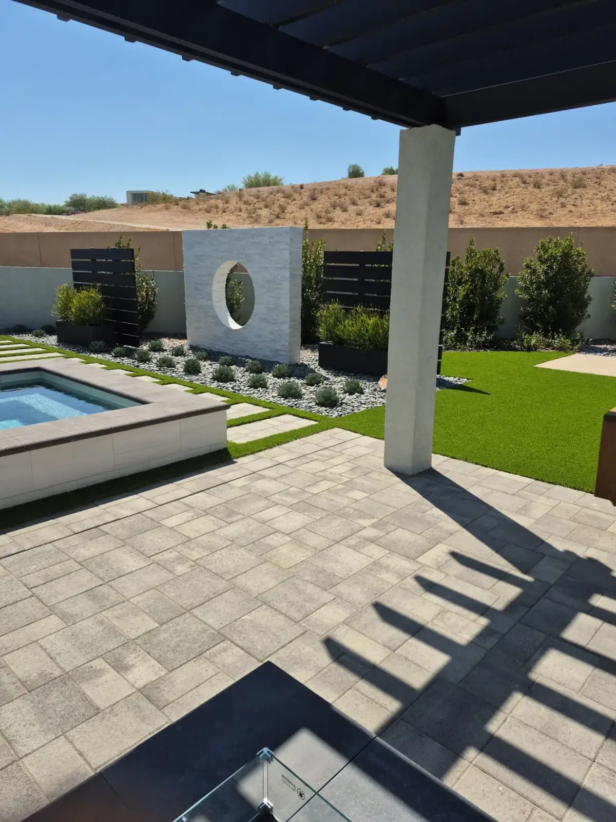 Modern hardscape patio and landscape design featuring a concrete pergola, raised pool, artificial turf lawn, decorative wall sculpture, and mixed plantings in a residential desert setting.