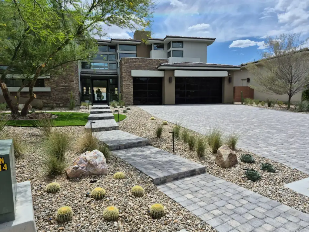 Desert xeriscaping and hardscape patio design featuring ornamental grasses, cacti, boulders, and pavers in a modern residential front yard.