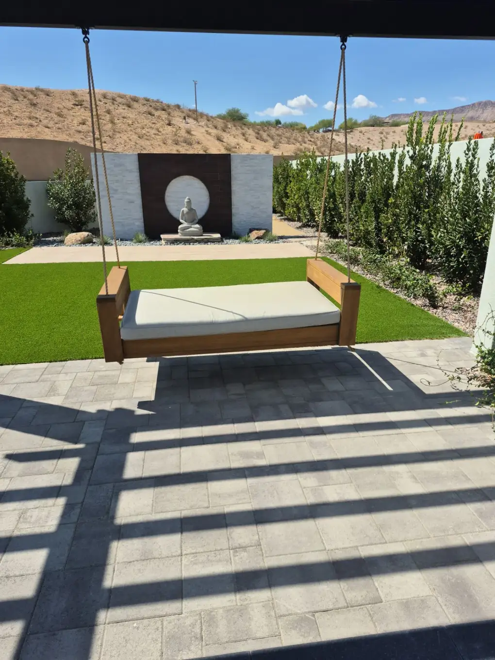A hardscape patio with integrated swing bed in a modern residential backyard landscape, featuring manicured lawn, privacy hedges, and a zen meditation feature wall with buddha statue.