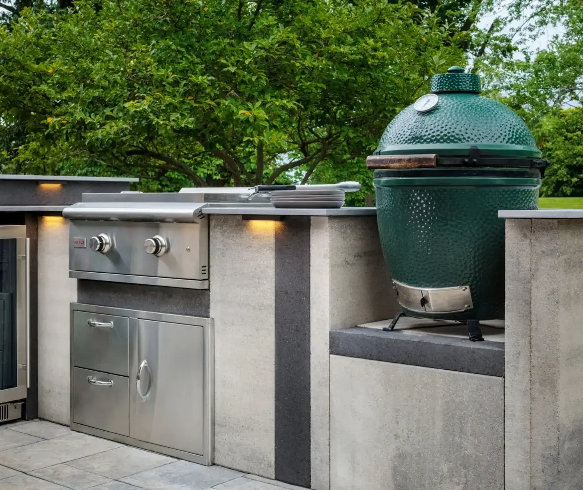 An outdoor kitchen and entertaining area featuring a built-in grill, storage cabinetry, and a green ceramic kamado-style cooker on a concrete patio, surrounded by mature trees.