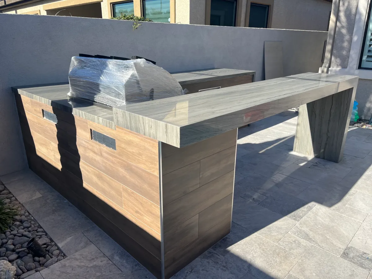 Custom outdoor kitchen with wood cabinetry and stone countertop in a residential patio setting.