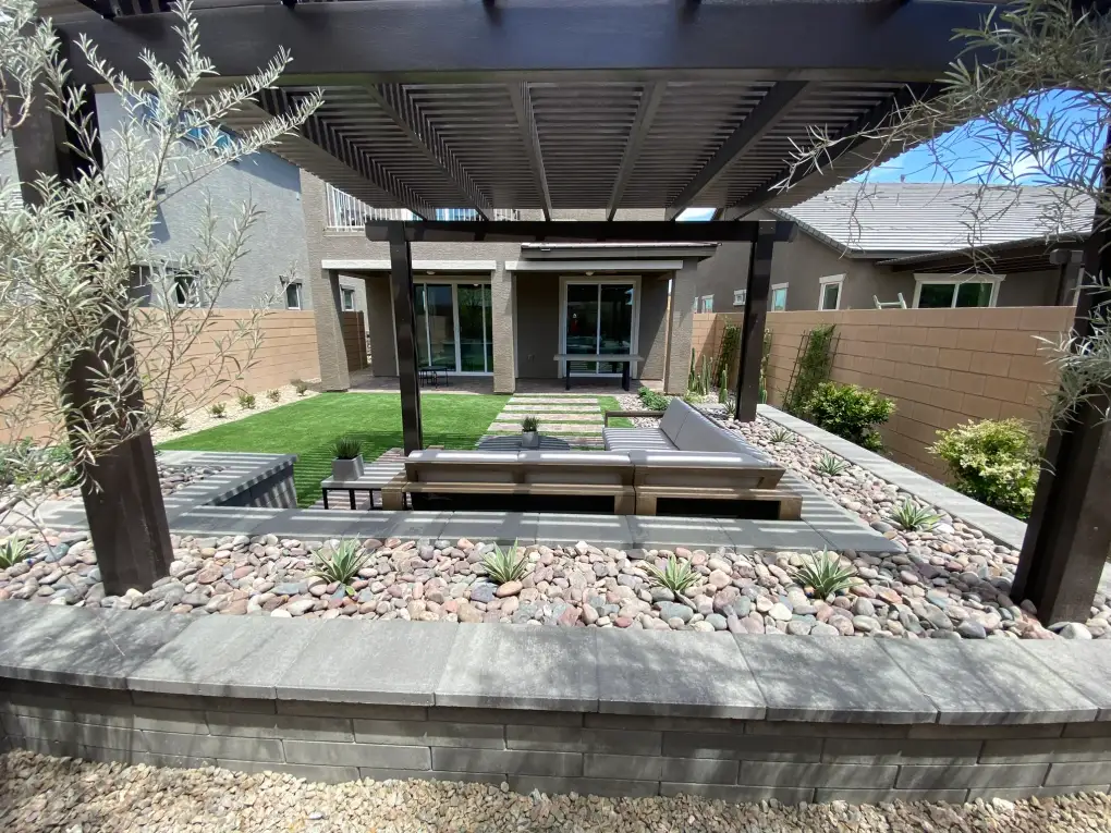 A modern hardscape patio with built-in seating and fire pit in a contemporary residential backyard, featuring composite decking, decorative river rock, and a pergola overhead.