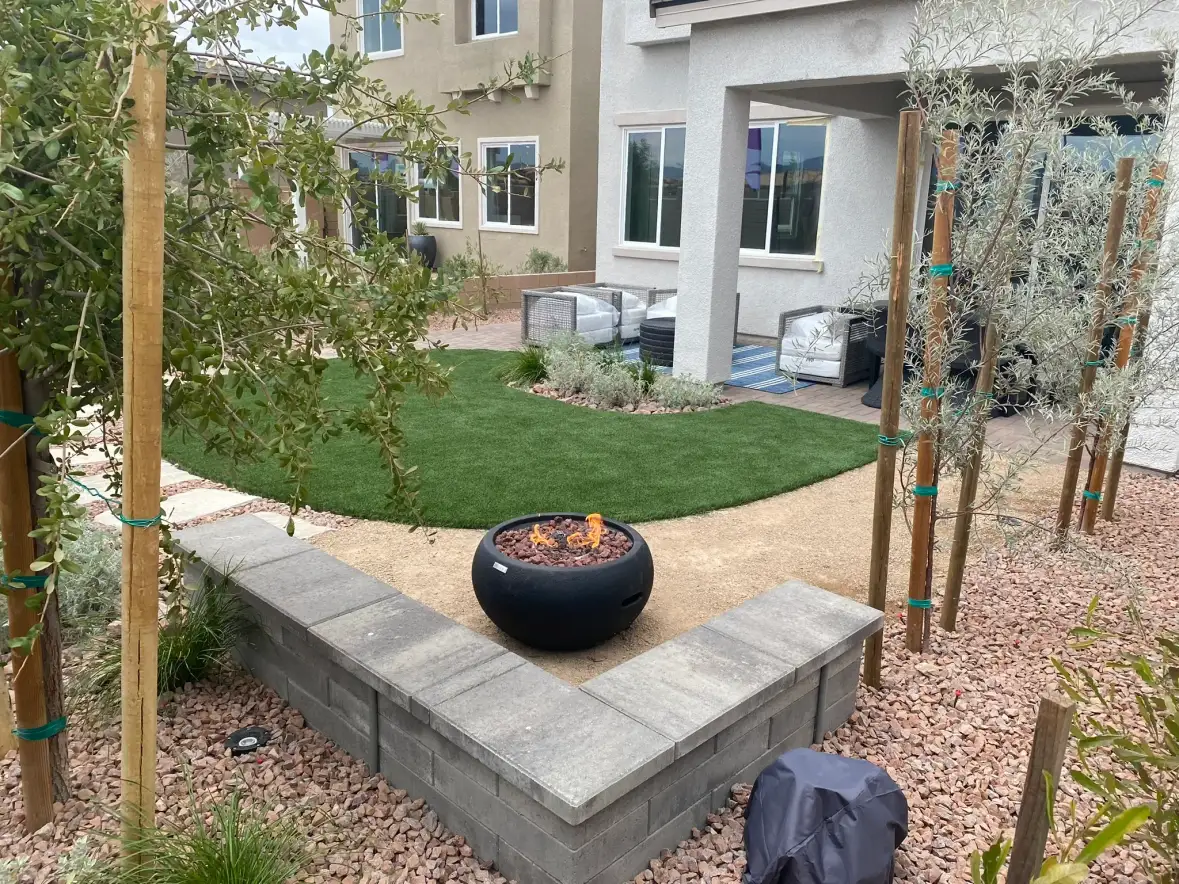 A modern backyard hardscape and landscape design featuring a concrete bench seating area with an integrated fire pit bowl, surrounded by gravel, young trees with support stakes, olive plants, and a manicured lawn with patio furniture in the background.