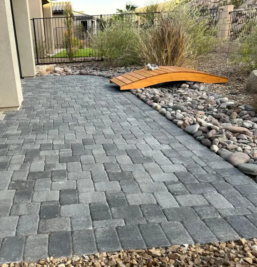 Paver patio installation with decorative wooden bridge and river rock edging in a residential backyard setting.