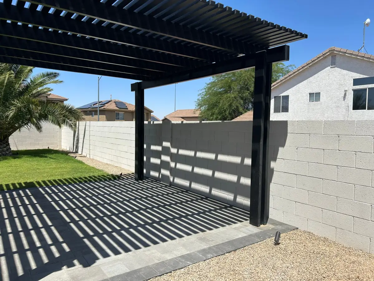 A black pergola structure with louvered roof panels installed over a paved patio area in a residential backyard setting.