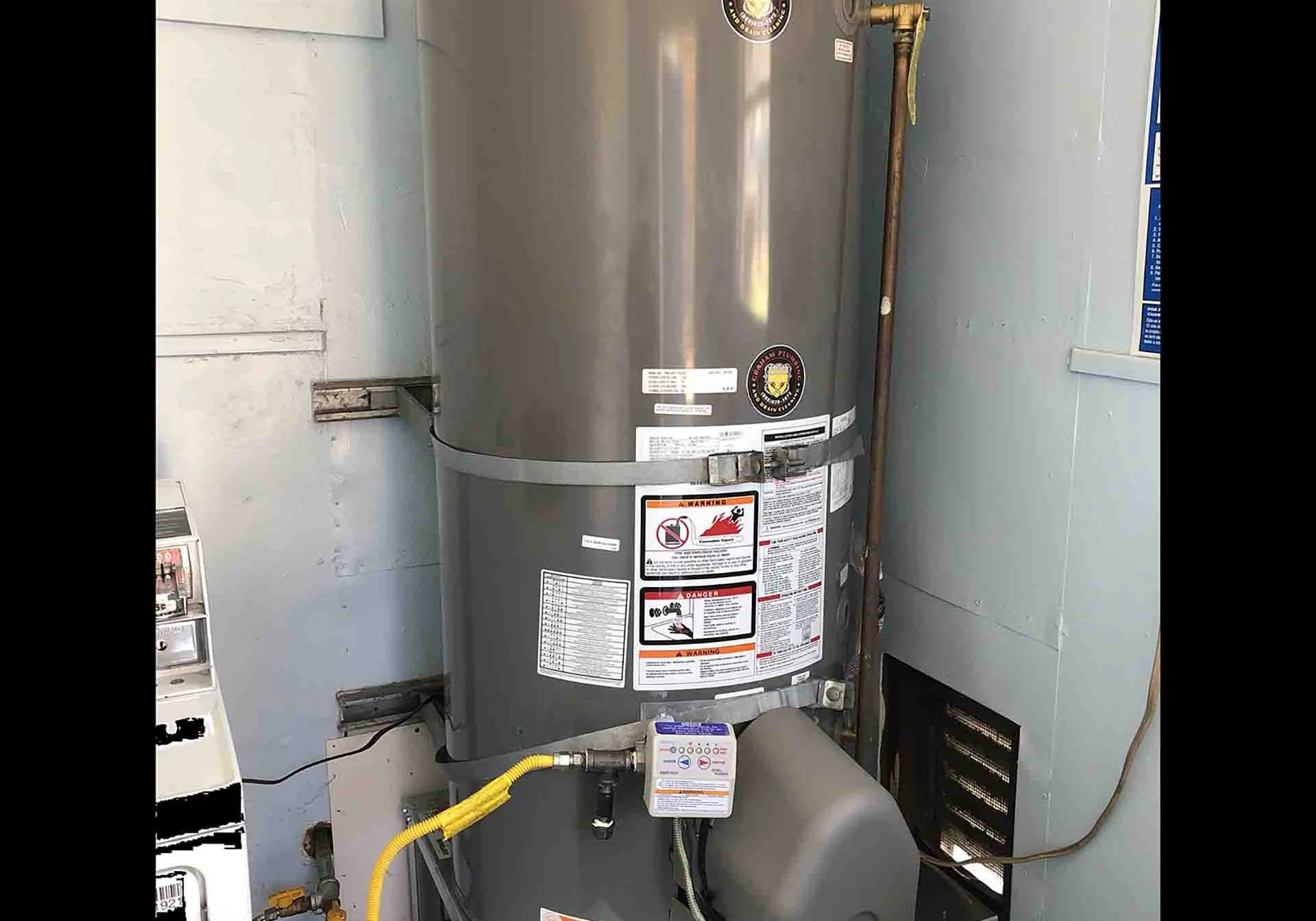 A tankless water heater unit installed in an indoor utility space with warning labels and gas connection visible.
