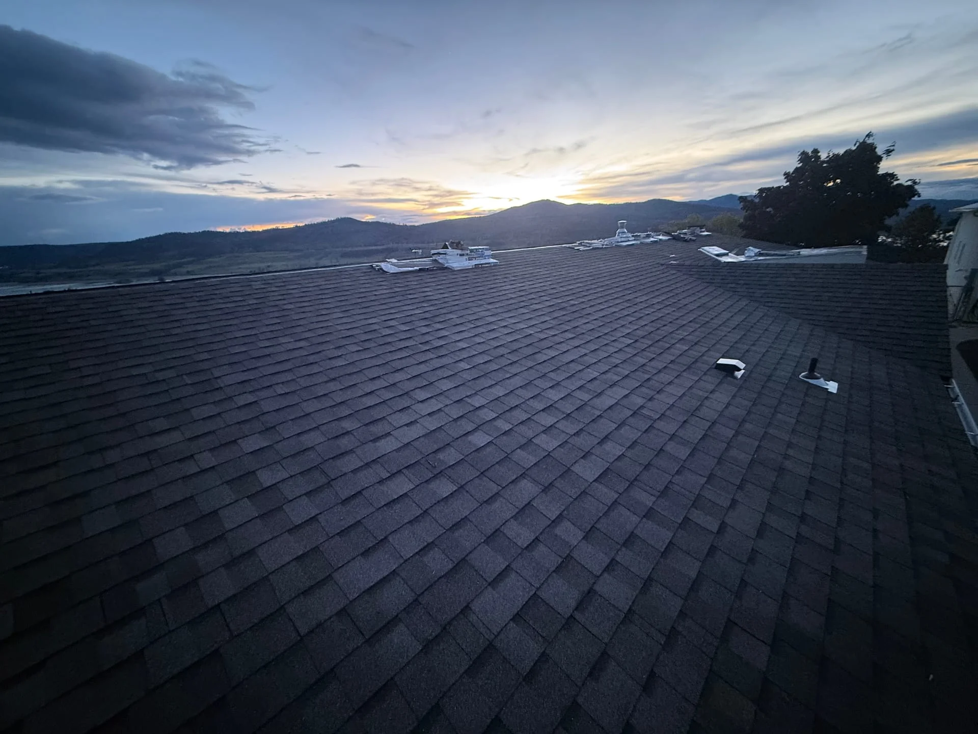 Asphalt shingle roofing installation on a residential roof at dusk with mountain valley backdrop.
