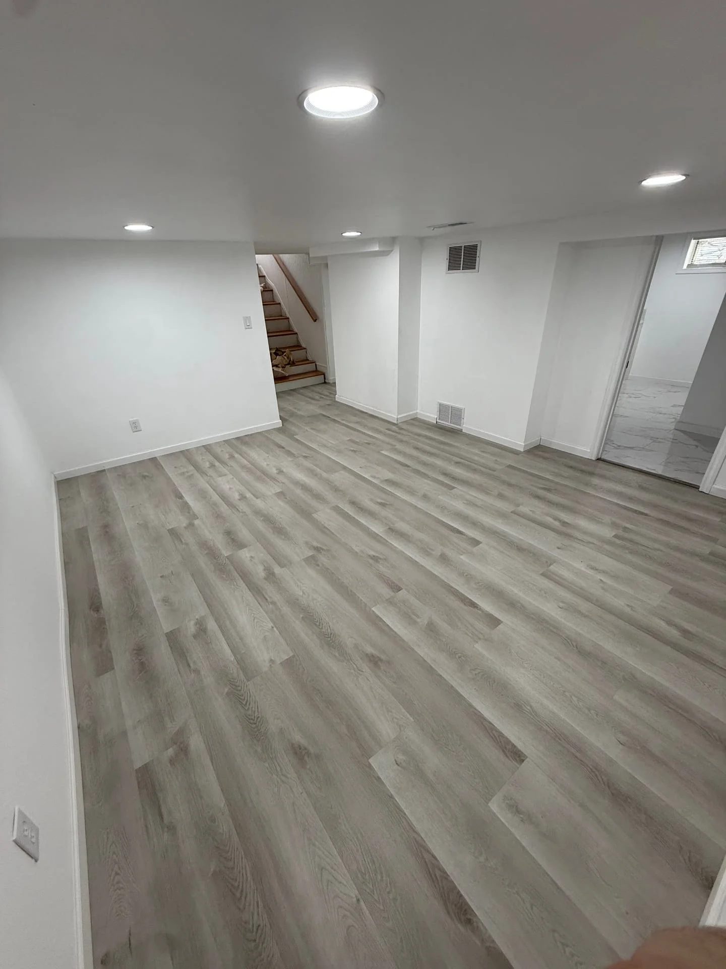 Vinyl plank flooring installation in a finished basement space with recessed lighting and white walls.