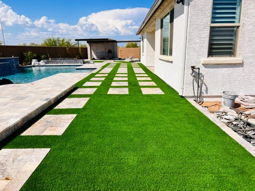 Artificial turf sod installation with stone steppers in a residential backyard alongside a pool and patio area.