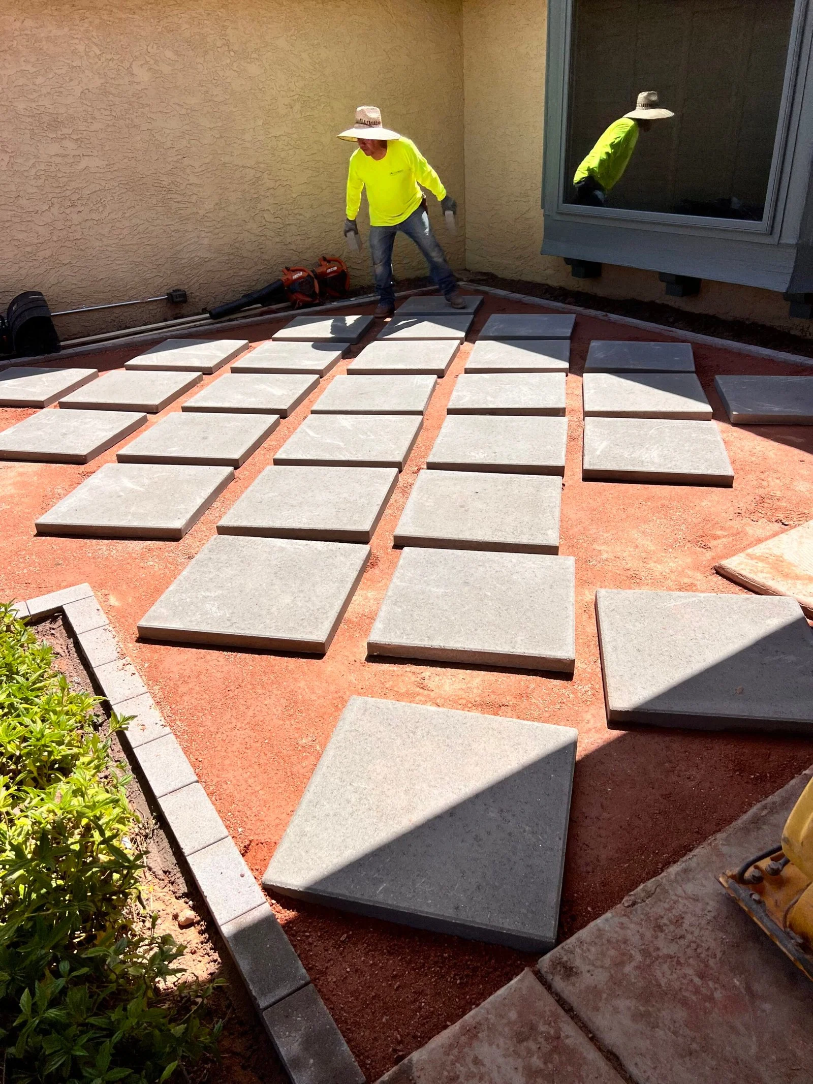 Concrete pavers being installed in a residential courtyard with reddish-brown base material and metal edging.