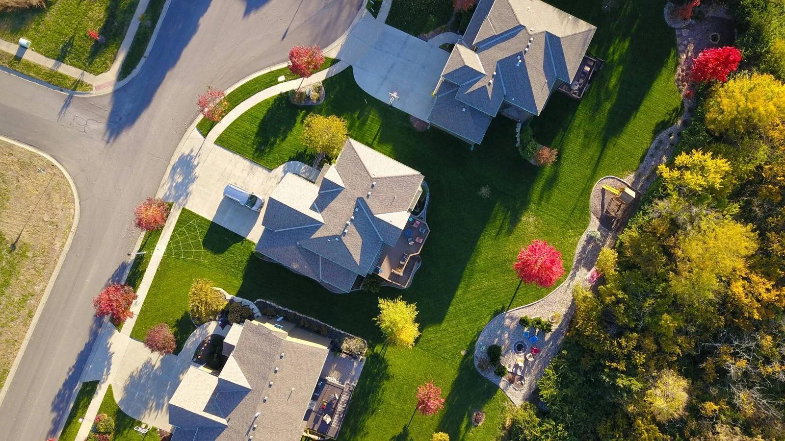 Residential lawn care and landscape maintenance in a suburban neighborhood aerial view, showing manicured lawns, ornamental plantings, and seasonal tree colors across multiple properties.