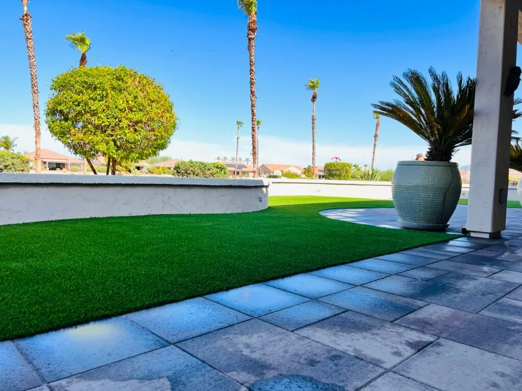 Synthetic turf lawn installation in a modern residential backyard with hardscape patio, palm trees, and decorative planter.