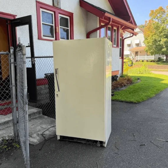 An old cream-colored refrigerator awaiting removal sits on a residential driveway next to a house.
