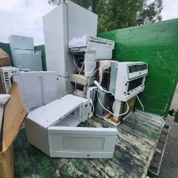 Discarded appliances and HVAC units stacked in a junk removal collection area with a green dumpster in the background.
