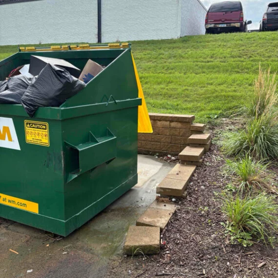 A green dumpster bin filled with bagged junk and debris in an outdoor residential setting with grass, landscaping, and parked vehicles in the background.