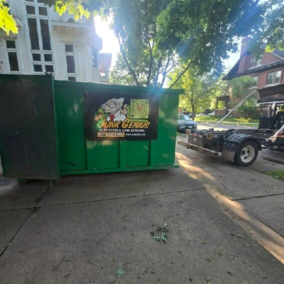 A green dumpster container for junk removal parked on a residential street lined with trees and buildings.
