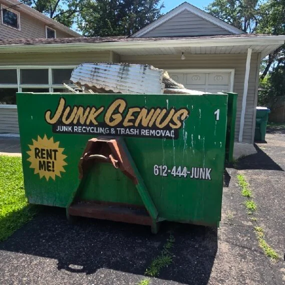 A green Junk Genius dumpster rental container positioned in a residential driveway in front of a house, displaying the company's branding and contact information.