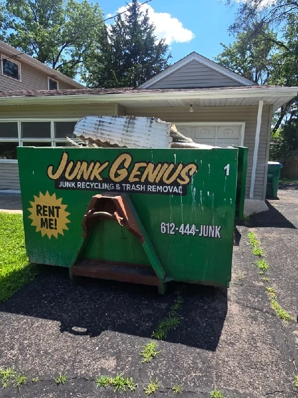 A green Junk Genius dumpster rental container filled with debris positioned in a residential driveway in front of a house.