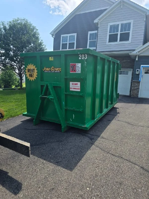 A green junk removal dumpster container positioned in a residential driveway next to a two-story house.