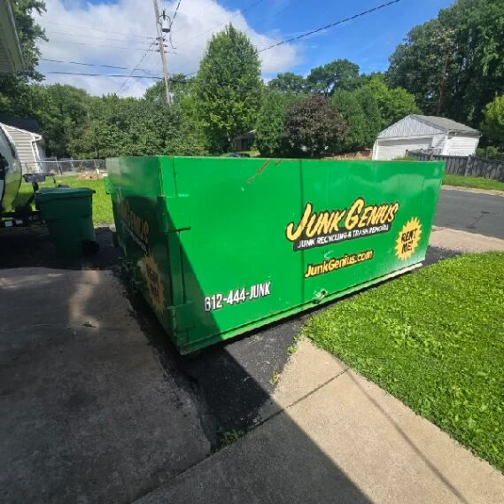 A green junk removal dumpster container positioned on a residential driveway in a suburban neighborhood.
