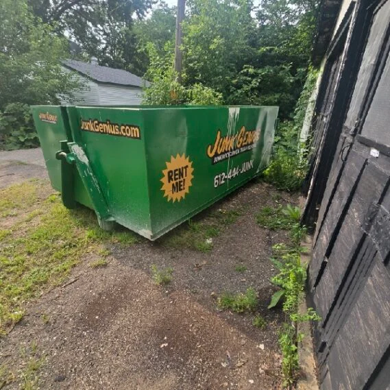 A green junk removal dumpster rental container positioned in a residential driveway next to a garage.