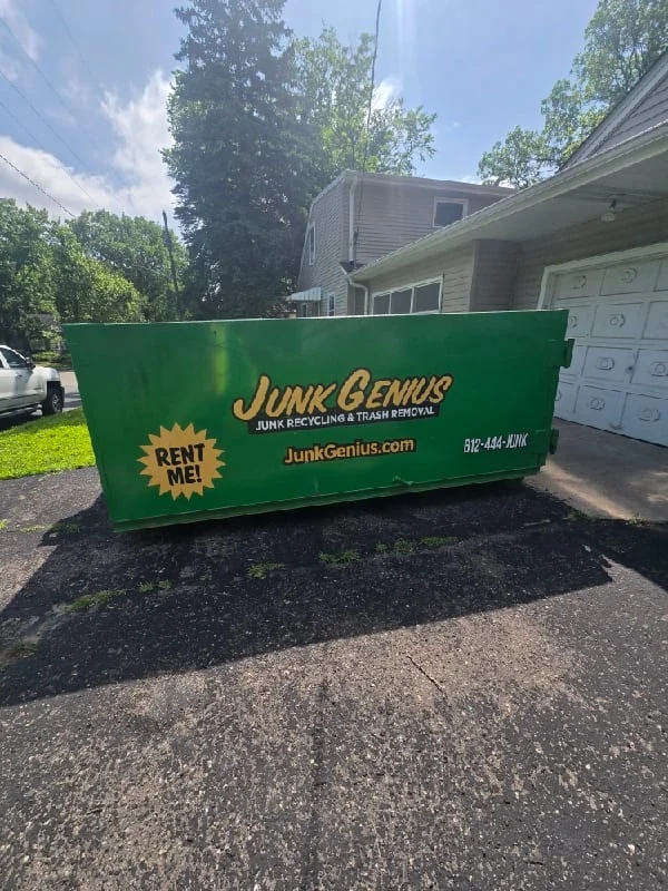 A green junk removal dumpster rental container positioned in a residential driveway next to a two-car garage.