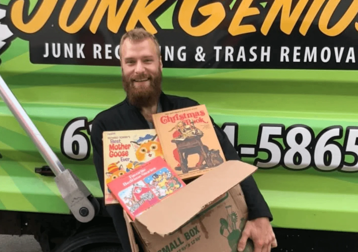 A junk removal employee holding a box of collected items including vintage books and holiday decorations in front of a branded junk removal truck.
