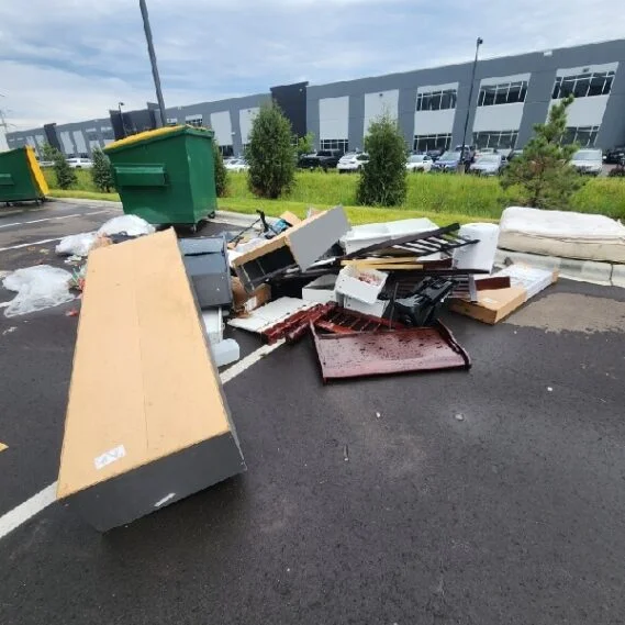 Junk removal service in progress at an industrial lot, showing a large pile of assorted debris including furniture, appliances, wood, and miscellaneous waste materials ready for disposal.