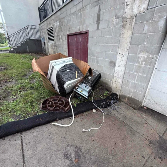 Junk removal service in progress at an exterior building foundation, showing accumulated items including appliances, metal parts, and debris being cleared from the site.