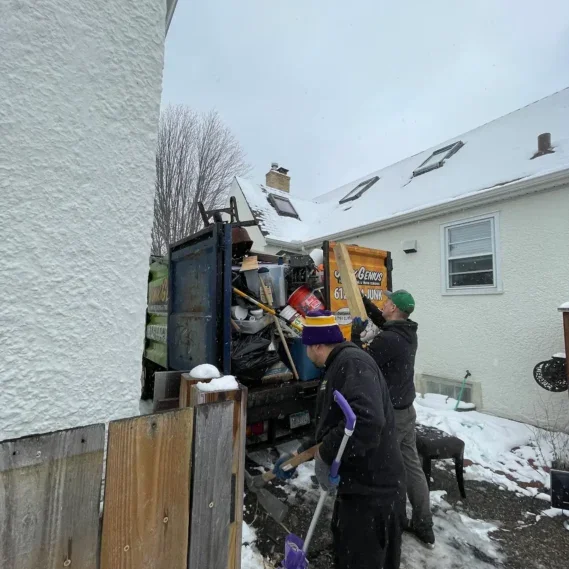 Junk removal service in progress at a residential property, with workers loading debris and waste materials into a large truck in a snowy backyard setting.