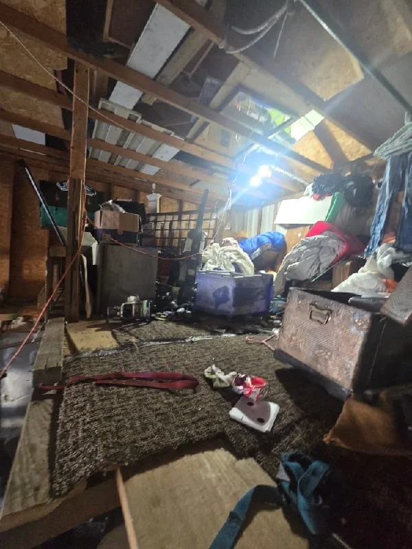 Junk removal service in progress inside a cluttered attic or storage space filled with accumulated items, boxes, and debris.