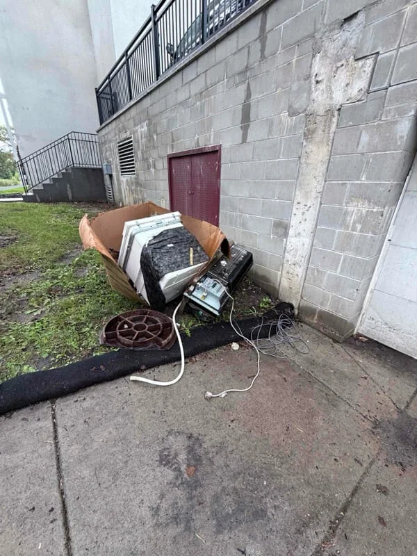 Junk removal service in progress showing discarded appliances and electronics in a cardboard box against a concrete building exterior.