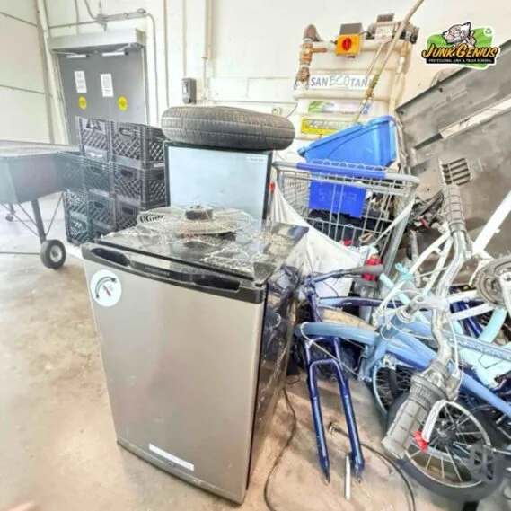 Junk removal service in progress showing a residential garage with appliances, furniture, bicycles, and miscellaneous items collected for hauling.
