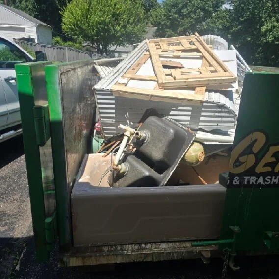 Junk removal service in progress, showing a loaded dumpster trailer filled with miscellaneous household waste, construction debris, and discarded items in a residential driveway.