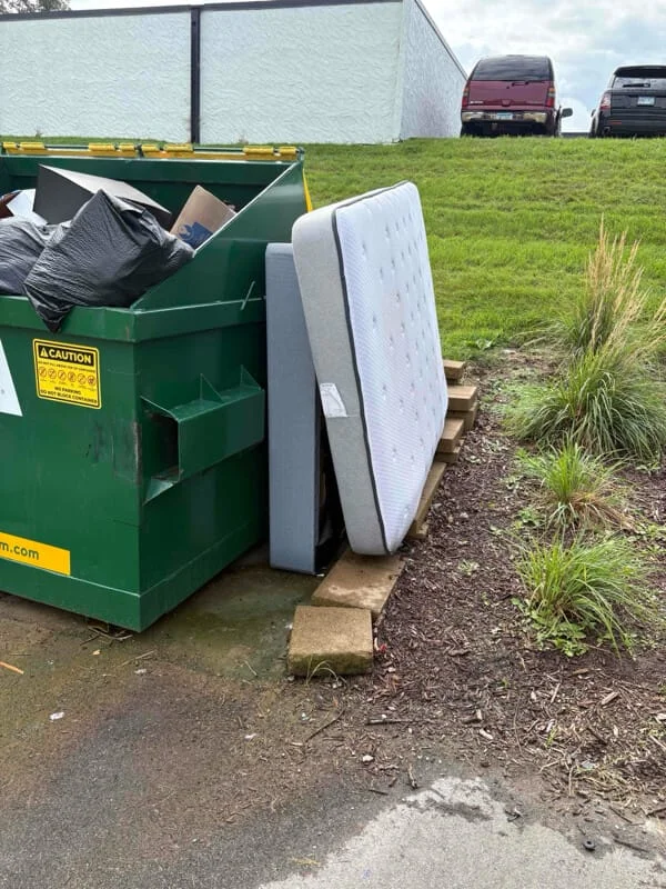 Junk removal service in progress showing mattresses and bags of debris being loaded into a green dumpster bin at a residential outdoor location.