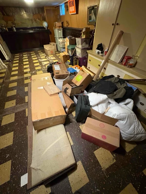 Junk removal service in-progress showing cluttered basement or garage space filled with cardboard boxes, clothing, and miscellaneous items awaiting removal.