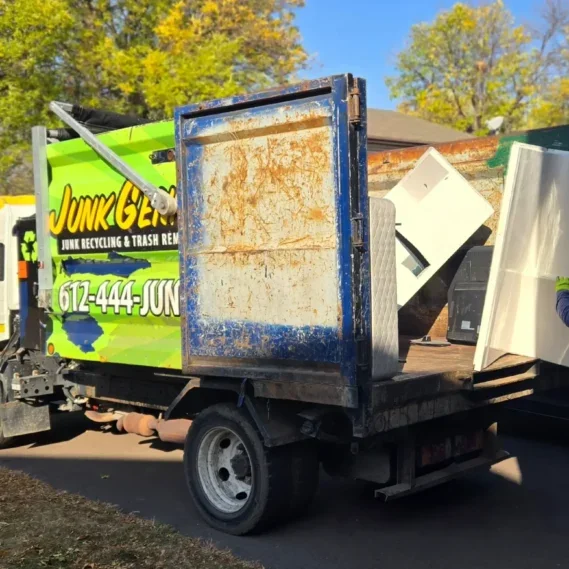 A junk removal truck with an open cargo bed containing discarded items and appliances, parked in a residential driveway during daytime.