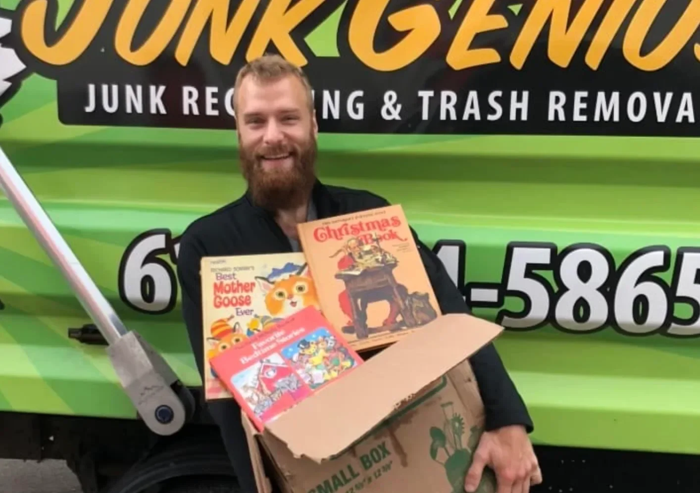 A junk removal worker holding a box of collected items including books and games in front of a green Junk Genius truck.