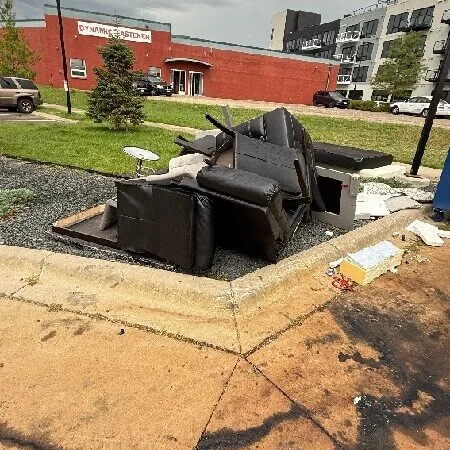 Large metal dumpster bin filled with black junk and debris in an outdoor urban lot next to a red brick building.