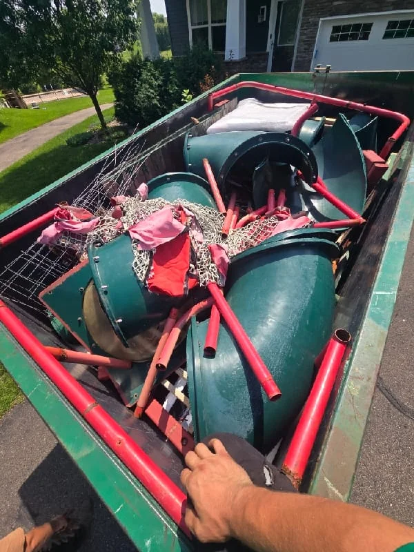 A loaded junk removal truck bed filled with discarded items including large green tanks, red piping, and miscellaneous debris in a residential driveway.