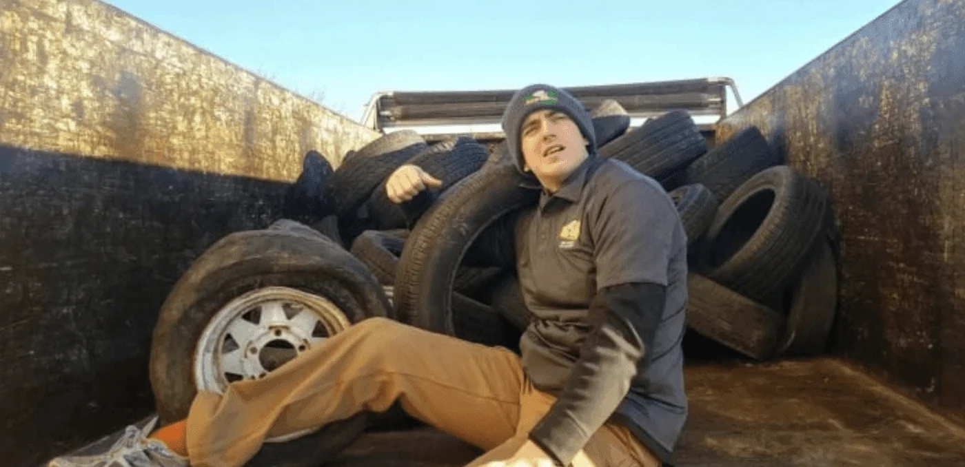 Tire removal and recycling work in progress at a junk yard with a worker posing among stacked used tires on a truck bed.