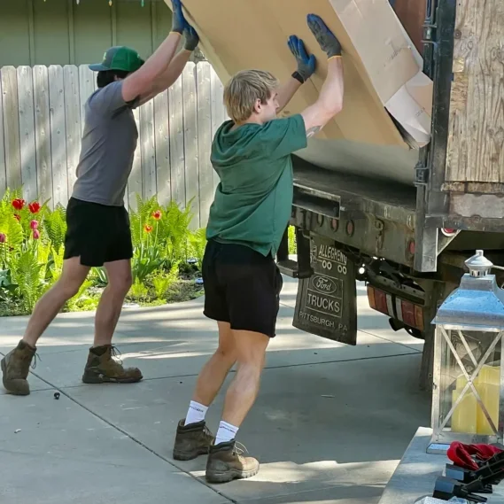 Two workers applying protective covering to a truck bed during a junk removal job in a residential driveway.