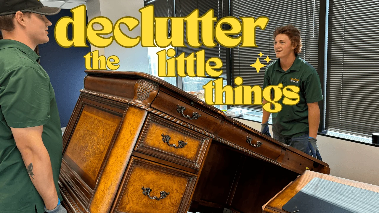 A wooden dresser being removed from an office space, with two employees in green uniforms standing beside it in front of a "declutter the little things" branded wall.